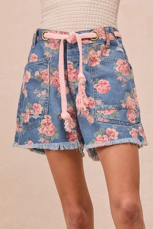 Close-up of floral denim shorts featuring a pink rose print, large front patch pockets, frayed hem, and a pink braided rope belt, styled at Sloan’s Formal Boutique.