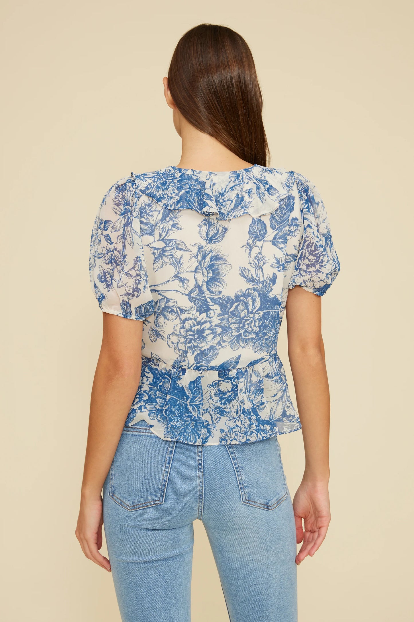 Back view of a woman wearing the Bee Ruffle Top featuring a blue floral print, short bubble sleeves, a subtle ruffle collar detail, and a gently cinched waist, paired with high-rise light wash jeans against a neutral background.