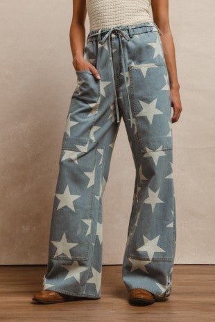 Front view of a model wearing wide-leg denim jeans with an ivory star print, drawstring waistband, and oversized patch pockets, styled with a textured sleeveless top for Sloan's Boutique.