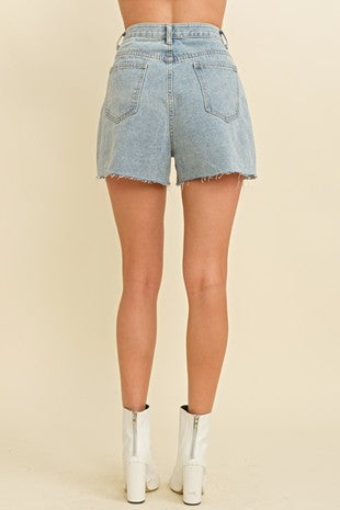 Back view of a woman wearing high-rise light-wash denim shorts with raw hems and back pockets, styled with white heeled ankle boots at Sloan’s Boutique.