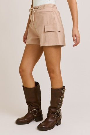 Side view of a woman wearing high-rise blush shorts with a drawstring waist and flap pocket detail, styled with knee-high brown buckle boots at Sloan’s Boutique.