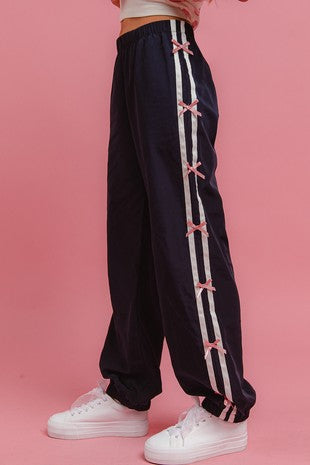 Side view of a model wearing navy track pants with white side stripes and pink bow accents, paired with white platform sneakers, styled against a pink background for Sloan’s Boutique.