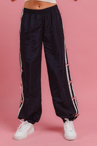 Front view of a model wearing navy track pants with pink bows and white side stripes, styled with white sneakers against a pink background for Sloan’s Boutique.
