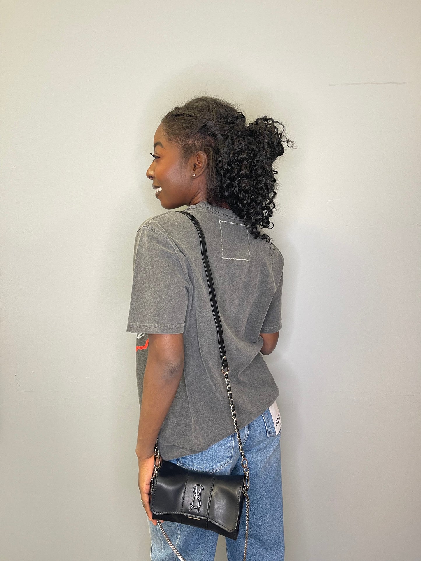 Back view of model wearing the tee, showing the faded charcoal fabric and square stitched patch, paired with light denim and a crossbody bag.