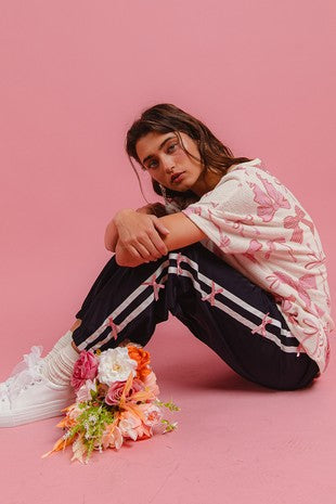 Woman sitting against a pink background wearing navy track pants with pink bow details and white side stripes, paired with a cream and pink bow-print top and white sneakers, styled for Sloan’s Boutique.
