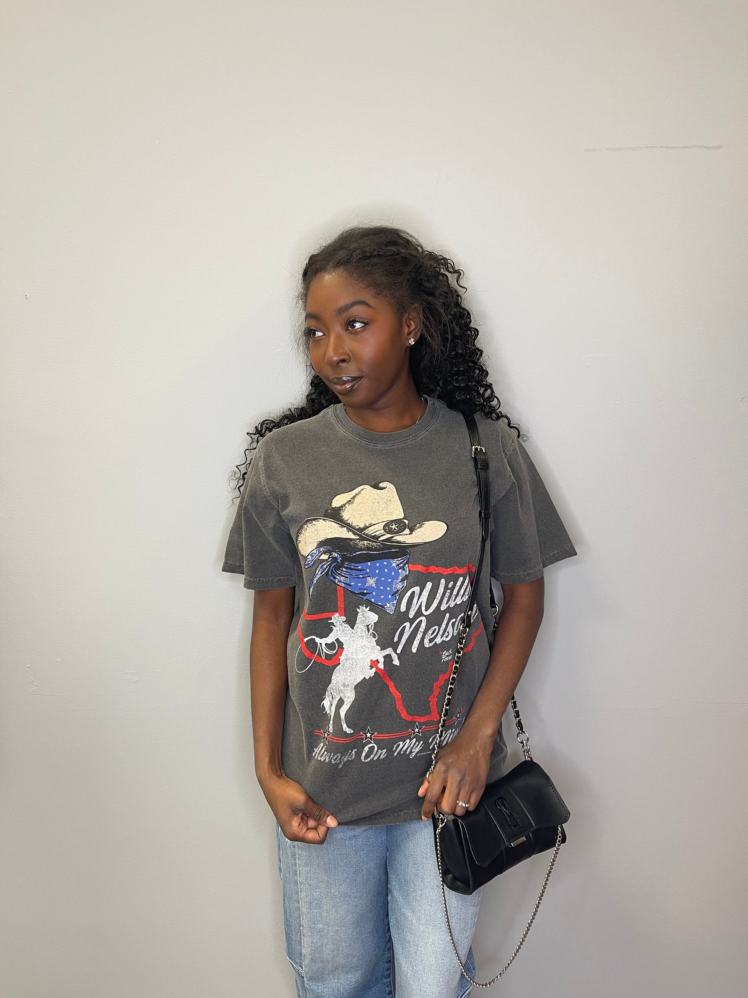 Model glances off to the side with a calm expression, displaying the front of the Willie Nelson tee with a black purse over the shoulder.