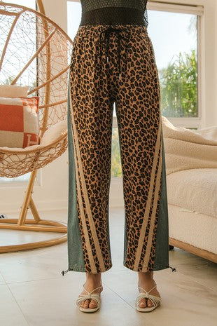Full front view of wide-leg leopard print pants with denim side panels, tan racing stripes, and drawstring waist, styled with strappy heels at Sloan’s Formal Boutique.