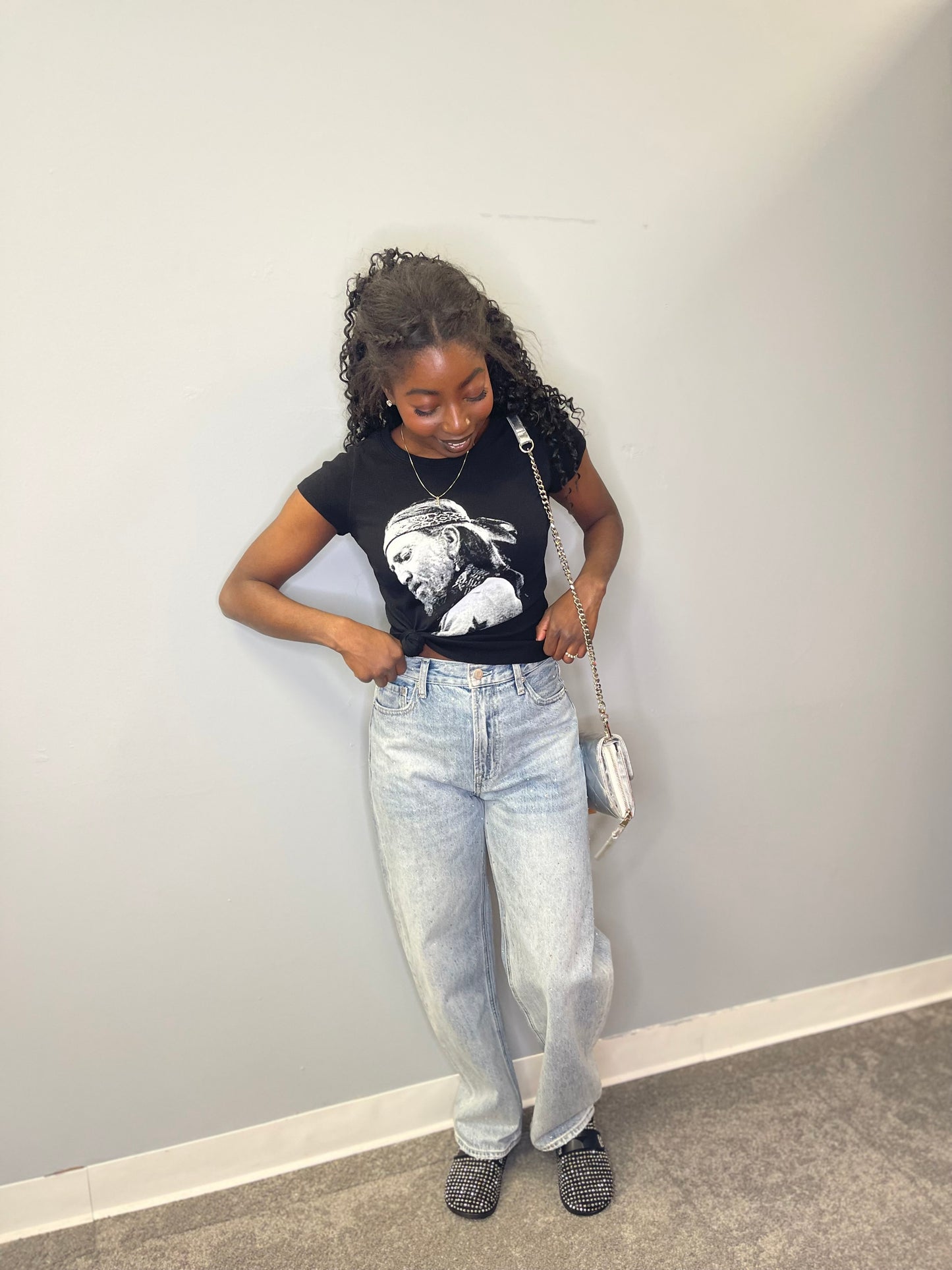 Full view of the model looking down and adjusting the tee, styled with light-wash jeans and studded black slip-on shoes.