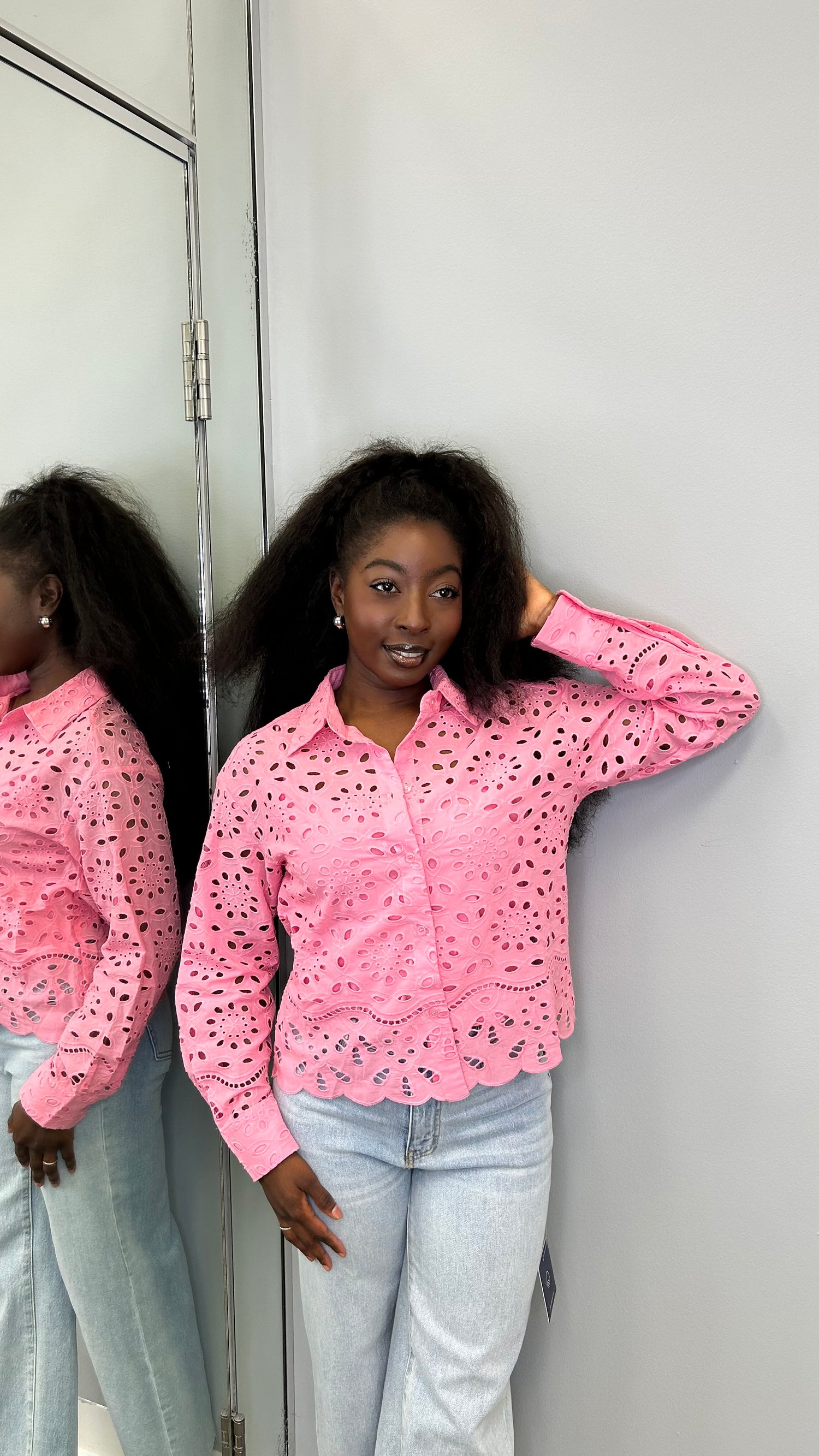 Woman wearing a bright pink floral eyelet blouse layered over a pink tank top, paired with jeans, smiling and posing in a boutique setting.