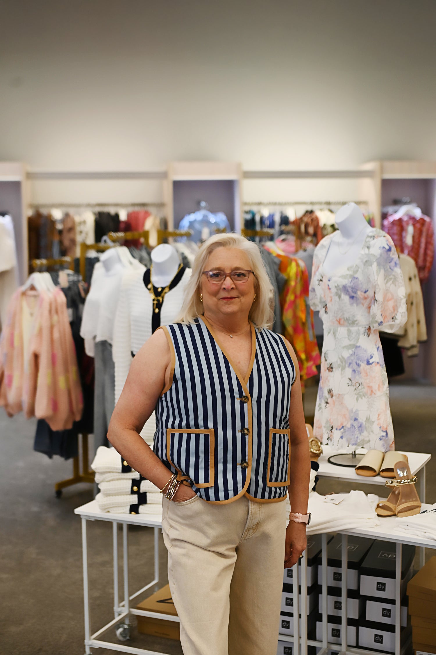 Woman wearing a sleeveless navy and white vertical stripe vest with camel brown trim and front buttons, paired with cream trousers in a boutique setting.