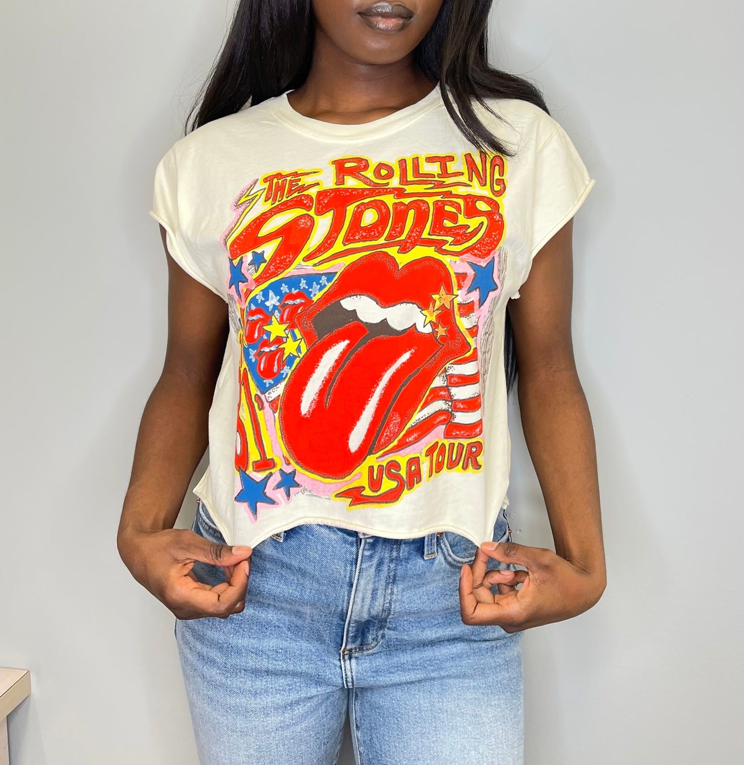 Front close-up of a model wearing the cropped Rolling Stones USA Tour tee, hands gently pulling the hem to show the raw edge and iconic red tongue logo with stars and stripes.