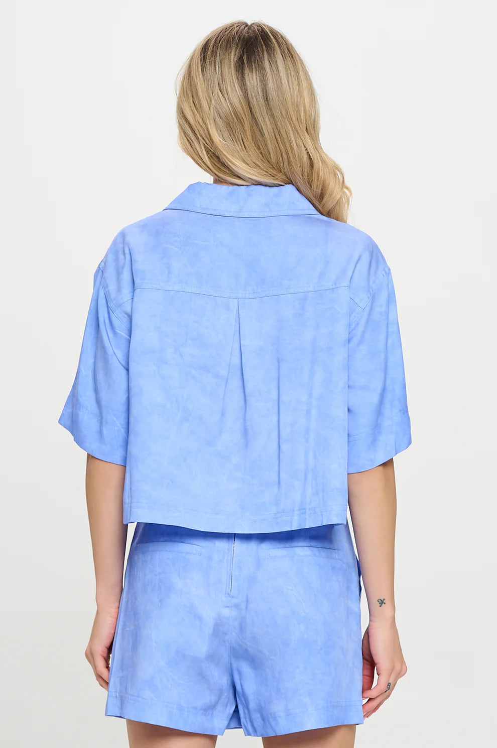 Back view of a woman wearing a sky-blue two-piece set featuring a short-sleeve, collared top with a back pleat and matching high-waisted shorts, styled with loose waves in her hair.