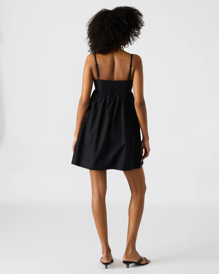 Back view of a woman wearing a black mini dress with thin adjustable straps and a smocked back panel, styled with black kitten heel sandals against a plain background.