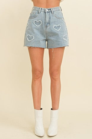 Front view of a woman wearing light-wash denim shorts with frayed hems and stitched heart designs, paired with white ankle boots at Sloan’s Boutique.