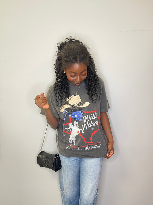 Model looks down while lightly pulling the hem of the Willie Nelson graphic tee, showing the full design with a crossbody bag and relaxed jeans.