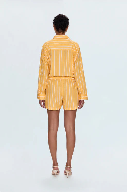 Back view of a woman wearing the Pistola Amelie Romper in yellow with white vertical stripes, featuring long sleeves, a collared neckline, and an elastic drawstring waist, styled with white heels at Sloan’s Formal Boutique.