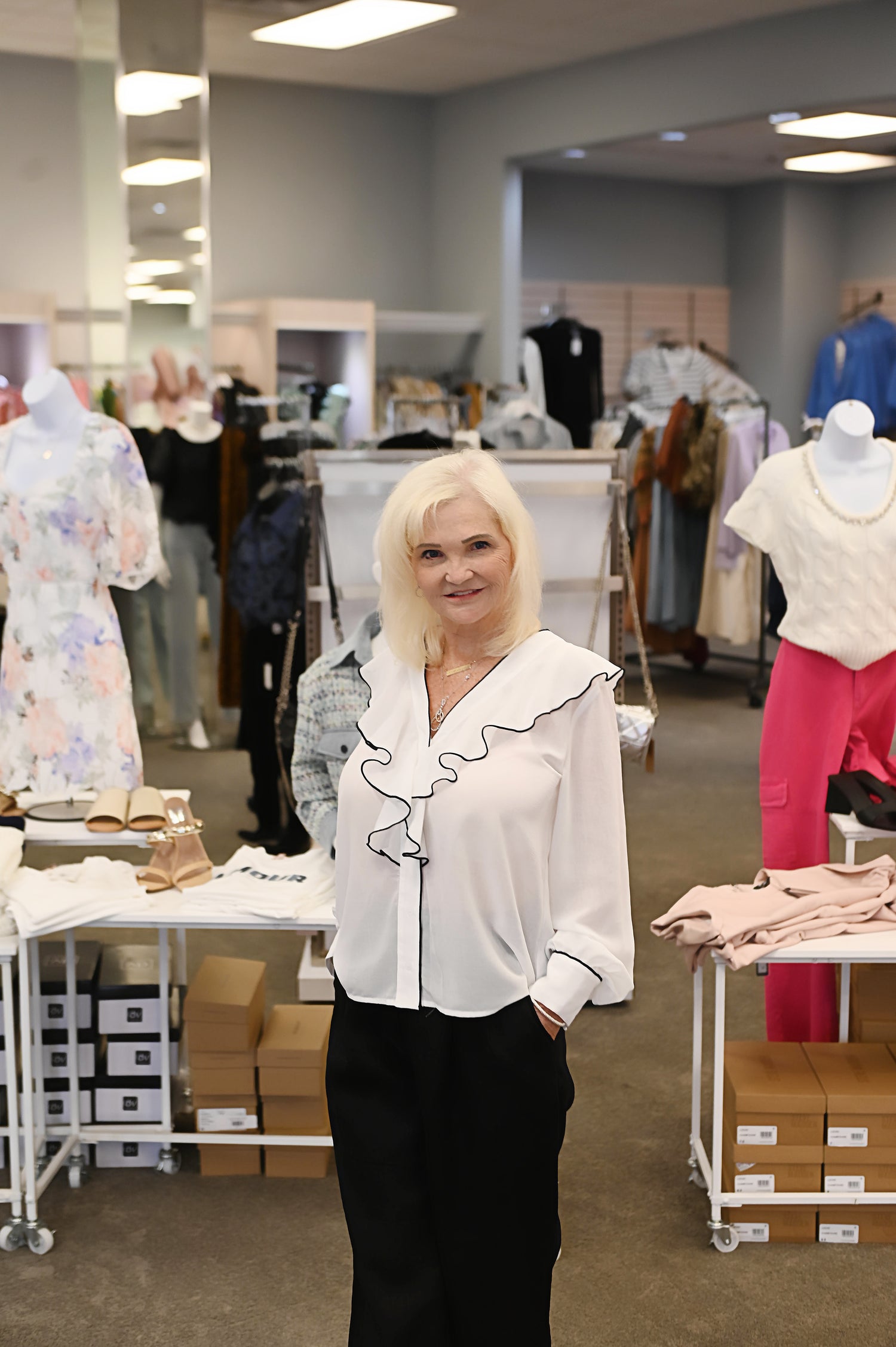 Woman wearing a white ruffle blouse and black pants, standing confidently inside a boutique with clothing displays in the background.