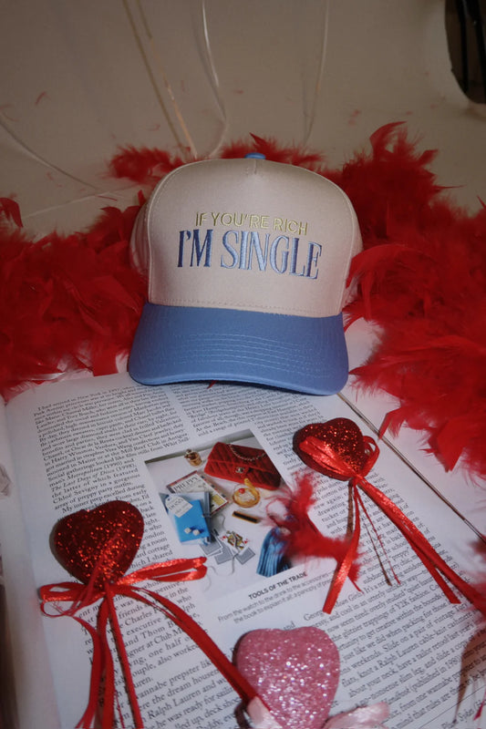 Cream and blue trucker hat with embroidered text that reads "If You're Rich I'm Single", styled on an open magazine with red heart decorations and red feather boas.