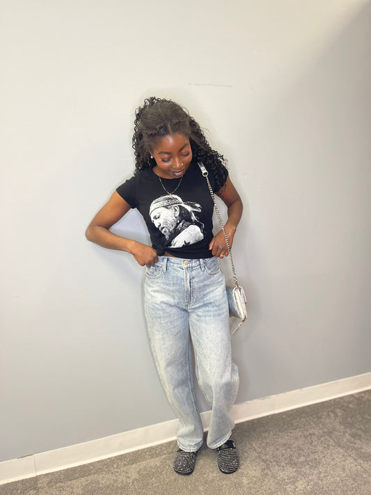 Full view of the model looking down and adjusting the tee, styled with light-wash jeans and studded black slip-on shoes.