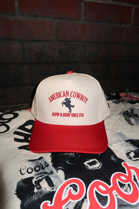 Cream and red trucker hat with “American Cowboy” embroidered above a bucking horse and the phrase “Boppin' & Ridin' Since 1776,” displayed on a vintage-style Coors print background.