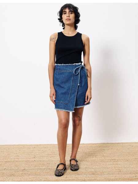 Front view of a woman modeling a wrap-style medium-wash denim mini skirt with a tie closure and raw edges, styled with a sleeveless black top.