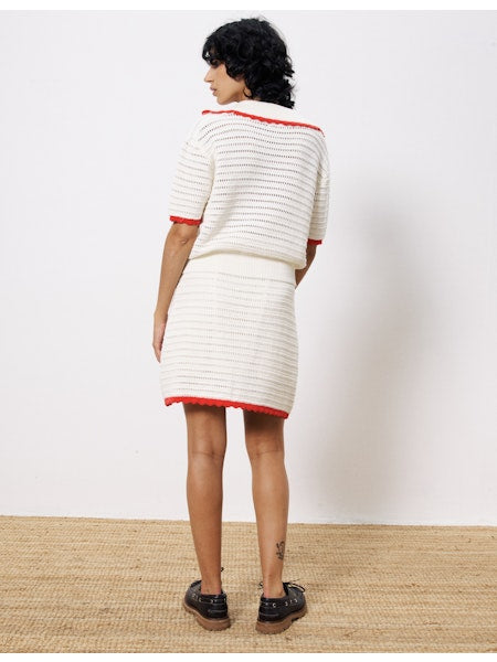Back view of a woman wearing an ivory crochet mini skirt with red scalloped trim, styled with a matching knit top and black shoes.
