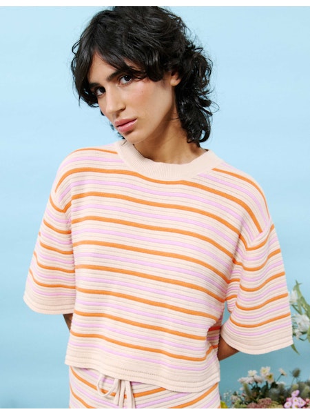 Model wearing the Anicia Top, a short-sleeved cotton sweater with horizontal stripes in pink, orange, yellow, and cream, styled against a pastel blue background with floral accents.