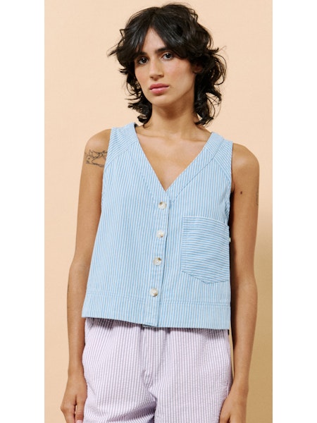 Model wearing the Colza Vest by FRNCH, a sleeveless blue and white striped vest with front buttons and a chest pocket, styled with lilac striped bottoms against a neutral peach background.