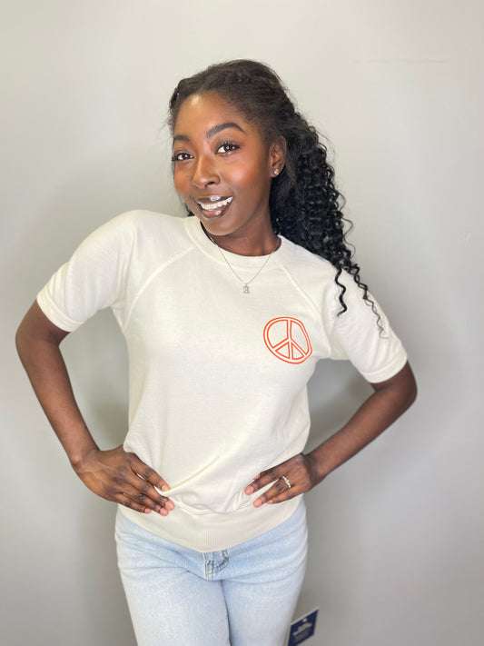 Model smiles with hands on hips, wearing a cream-colored short-sleeve raglan tee with an orange peace sign on the chest, paired with light denim.