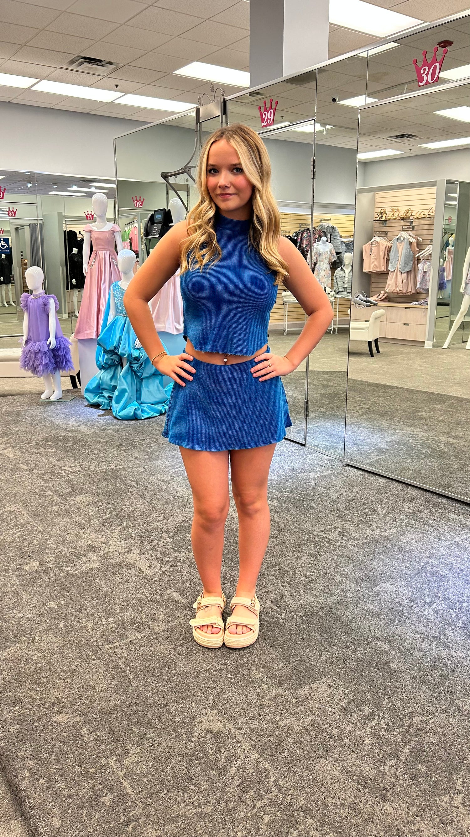 Woman wearing a blue two-piece set featuring a worn-detail tank top and matching mini skort, posing in front of mirrored doors.