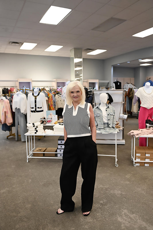 Woman wearing a sleeveless ivory sweater top with black vertical stripes and a split collar, paired with high-waisted black trousers, posing in a boutique setting.