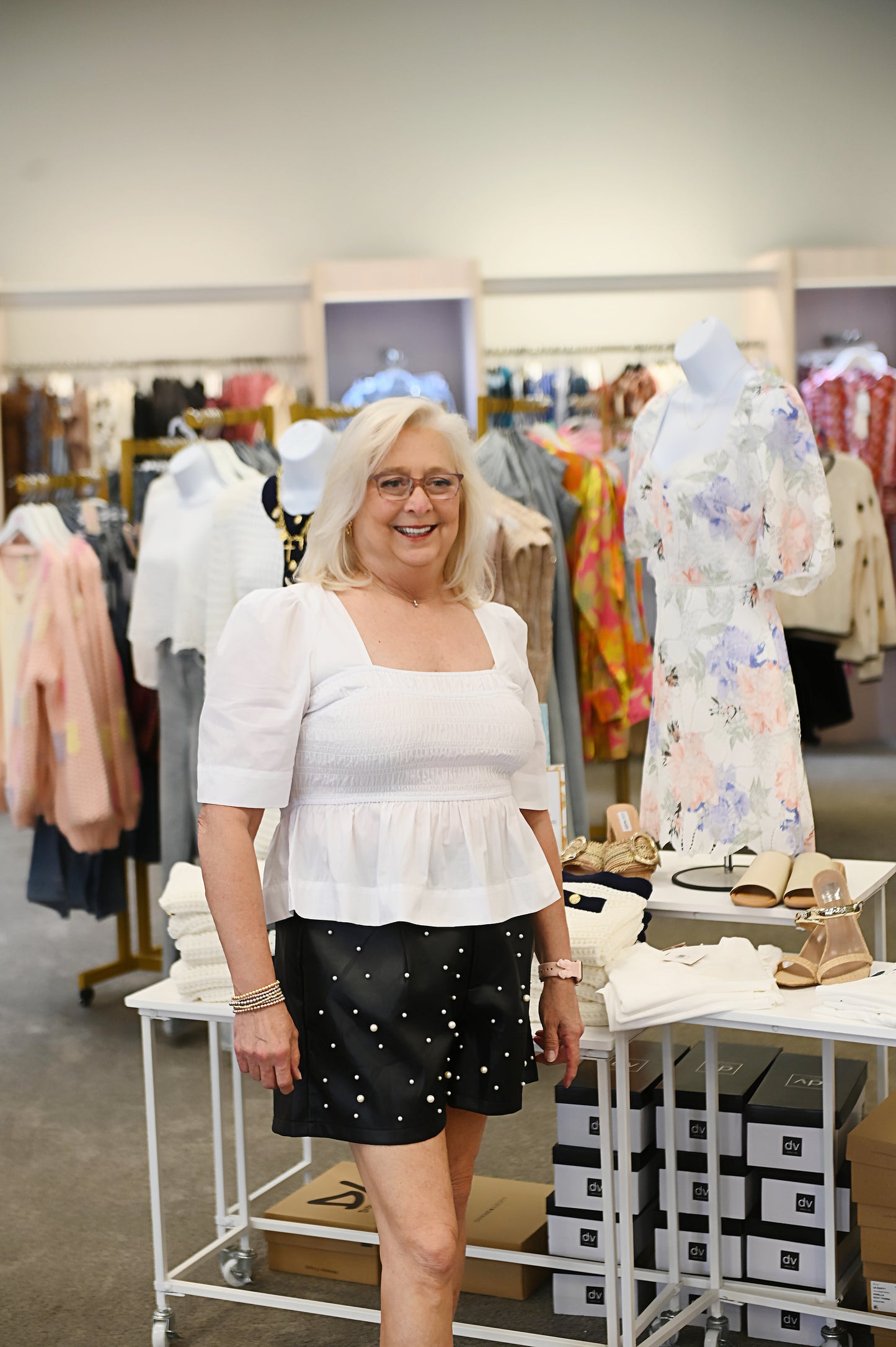 Woman wearing black high-waisted shorts with pearl embellishments and a flowy white smocked blouse, standing in a boutique beside a table of neutral-toned accessories.