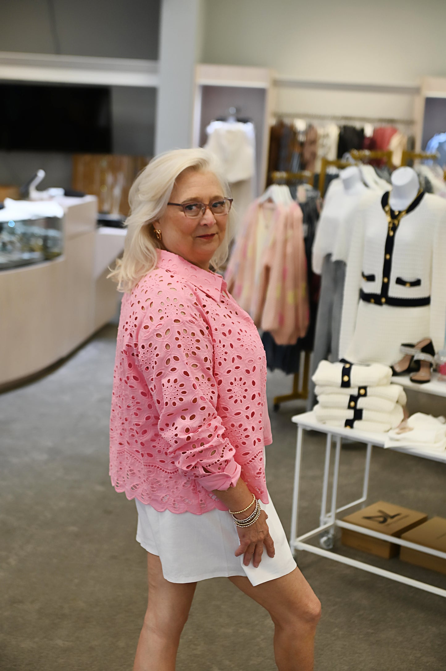 Woman wearing a bright pink floral eyelet blouse layered over a pink tank top, paired with a white skirt, smiling and posing in a boutique setting.