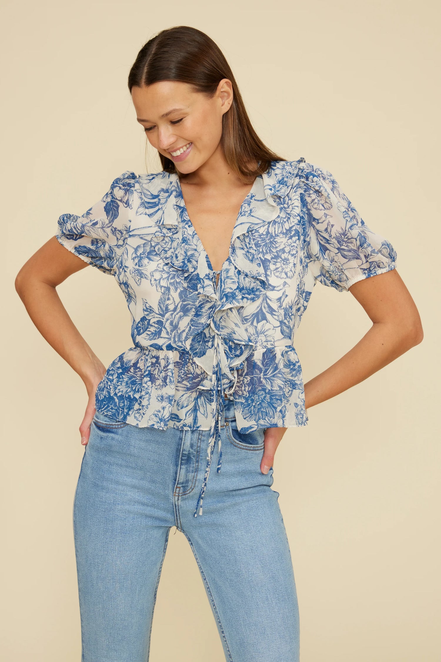 Front view of a woman wearing the Bee Ruffle Top in a blue floral print, featuring a ruffled V-neckline, short bubble sleeves, and a tie-front waist detail, paired with light-wash high-rise jeans against a neutral background.