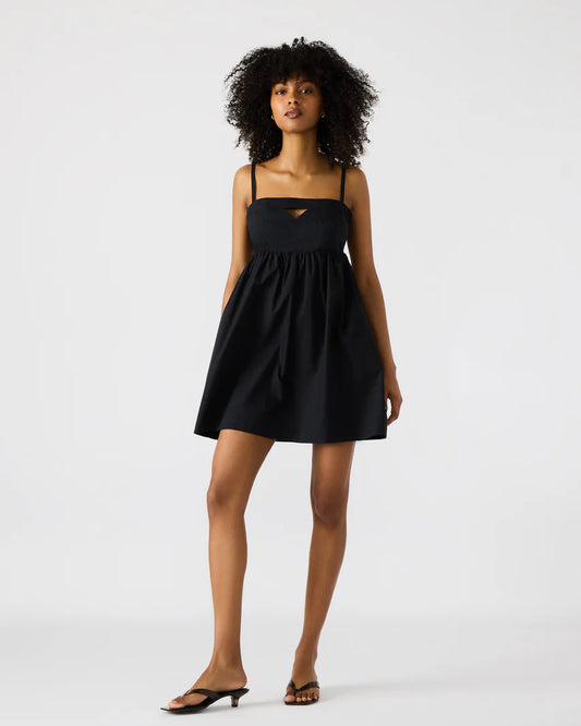 Woman wearing a black sleeveless mini dress with spaghetti straps, a sweetheart neckline with a small front cutout, and an A-line silhouette, styled with black heeled sandals against a light background.