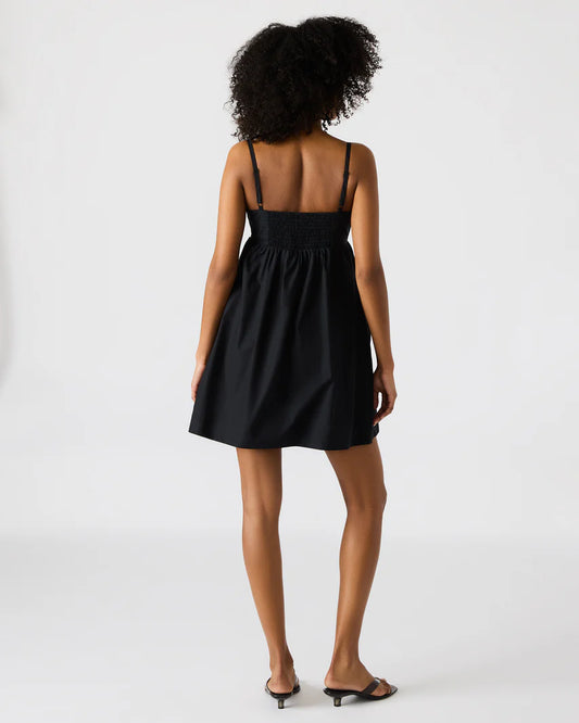  Back view of a woman wearing a black mini dress with thin adjustable straps and a smocked back panel, styled with black kitten heel sandals against a plain background.