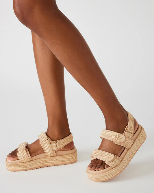 Close-up side view of a woman wearing Steve Madden BIGMONA platform sandals in natural raffia, showing the textured double straps and flatform sole against a white background.
