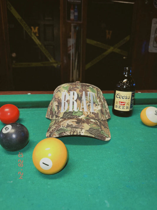 Camo print trucker hat with bold white “BRAT” embroidery, placed on a pool table surrounded by billiard balls and a vintage Coors beer bottle.